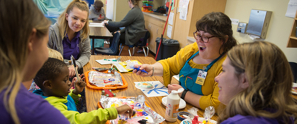 Student teachers are sitting around a table painting with a small child. 